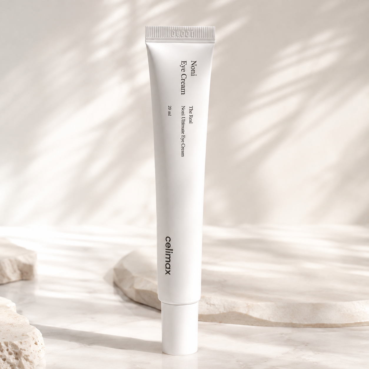 Celimax Noni Eye Cream tube standing on marble with soft dappled shadow, technology formula flat-lay